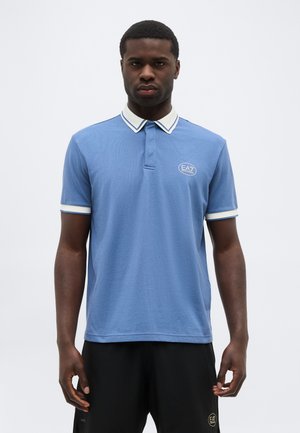 Άνδρας που φοράει μπλε κοντομάνικο πόλο μπλουζάκι με λευκή διακόσμηση στο γιακά και στα μανίκια, και μαύρο σορτς με μικρές λεπτομέρειες λογότυπου.