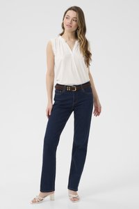 Haut blanc sans manches avec des épaules volantes, associé à un jean droit bleu foncé et une ceinture brune avec une boucle dorée. Sandales à talons blocs blanches.