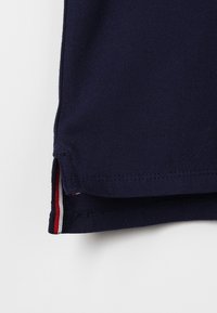 Granatowa koszulka polo z fakturowanej tkaniny, z bocznymi wentylami i kontrastującymi czerwono-białymi wykończeniami u dołu.