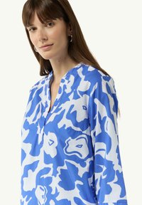 Blaue und weiße gemusterte Bluse mit Kragen, locker geschnitten und mit langen Ärmeln. Sichtbare Tasche auf der linken Seite. Baumwollmischgewebe.