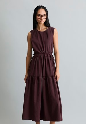 Vrouw met lang zwart haar en bril, gekleed in een donkerpaarse mouwloze midi-jurk met een geplooide taille en geplooide rok, staand tegen een grijze muur.