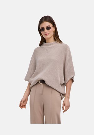 Femme portant un pull poncho en maille beige, un pantalon beige taille haute, une ceinture noire, des boucles d'oreilles créoles dorées, des lunettes de soleil et un bracelet doré, debout devant un fond blanc.