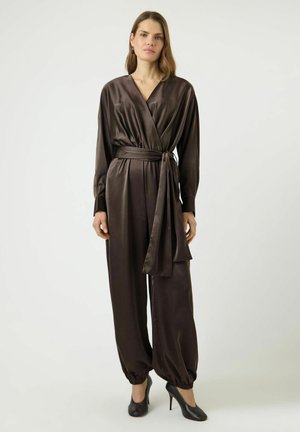 Kobieta ubrana w ciemnobrązowy satynowy kombinezon z długimi rękawami, z paskiem w talii, marszczonymi nogawkami i czarnymi czółenkami na wysokim obcasie, stojąca na jednolitym tle.