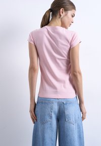 Γυναίκα με καστανά μαλλιά σε αλογοουρά, που φορά σκουλαρίκια, ένα εφαρμοστό ροζ κοντομάνικο μπλουζάκι και ανοιχτόχρωμα φαρδιά τζιν, στέκεται μπροστά σε έναν λευκό τοίχο.