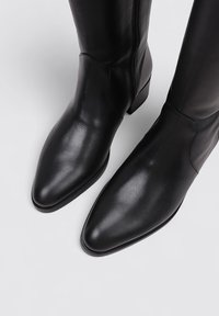Bottes noires en cuir jusqu'aux genoux avec un bout pointu et une fermeture éclair latérale, présentant une texture lisse et un talon bas rectangulaire.