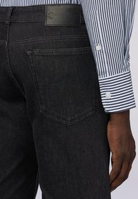 Jeans de mezclilla negra con un diseño estándar de cinco bolsillos, con una etiqueta de cuero texturizado en la cinturilla y un puño de camisa a rayas visible.
