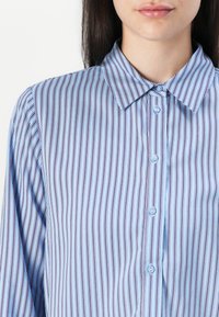 Camisa de manga longa azul claro com finas riscas verticais em roxo escuro. Apresenta um colarinho pontudo e parte frontal com botões azuis claros.