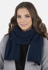 Sötétkék kötött sál a nyak köré drapírozva, puha textúrával és egyenes szélekkel, amelyek kiegészítik a szürke kabátot és a fekete felsőt.