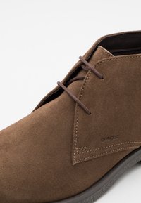 Brown suede ankle boot with a rounded toe, lace-up design, and visible stitching. Features a textured rubber sole and branding on the side.