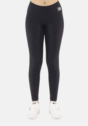 Vrouw draagt zwarte leggings met hoge taille met wit "LEONE" logo op de heup, gecombineerd met witte sneakers, staand tegen een effen lichte achtergrond.