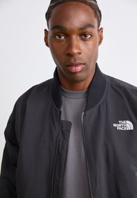 Veste aviateur noire avec fermeture éclair à l'avant, col côtelé et logo sur la poitrine gauche, portée sur un sweat-shirt gris. Texture de tissu lisse.