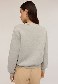 Felpa grigia con scollatura tonda, decorata con borchie lungo la schiena. Maniche lunghe e vestibilità comoda, realizzata in tessuto morbido.