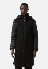 Sort pufferjakke med fleece-krop, der har trykknapper, to frontlommer og hætte med snor. Teksturerede ærmer giver kontrast.