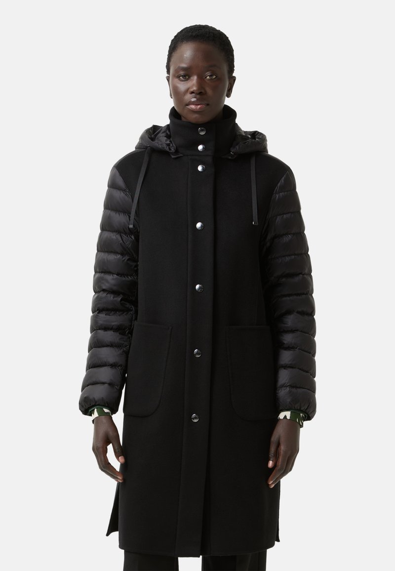 Sort pufferjakke med fleece-krop, der har trykknapper, to frontlommer og hætte med snor. Teksturerede ærmer giver kontrast.