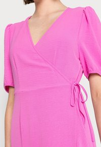 Vestido envolvente fucsia con un profundo escote en V, mangas cortas abullonadas y un lazo lateral. Tela suave y texturizada con una silueta fluida.