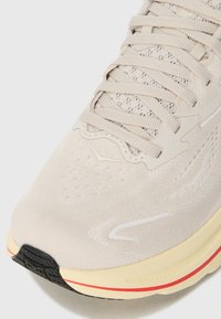 Chaussure de sport beige clair avec une tige en tissu texturé, des lacets plats et une semelle rembourrée. Présente une semelle extérieure noire et jaune proéminente.