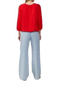 Blouse rouge à manches longues avec détails froncés au dos, associée à un jean en denim large et bleu clair. Texture lisse et coupe décontractée.