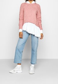Pull en maille rose avec un ourlet volanté blanc asymétrique, porté sur une chemise à col, associé à un jean droit bleu clair et des baskets blanches.