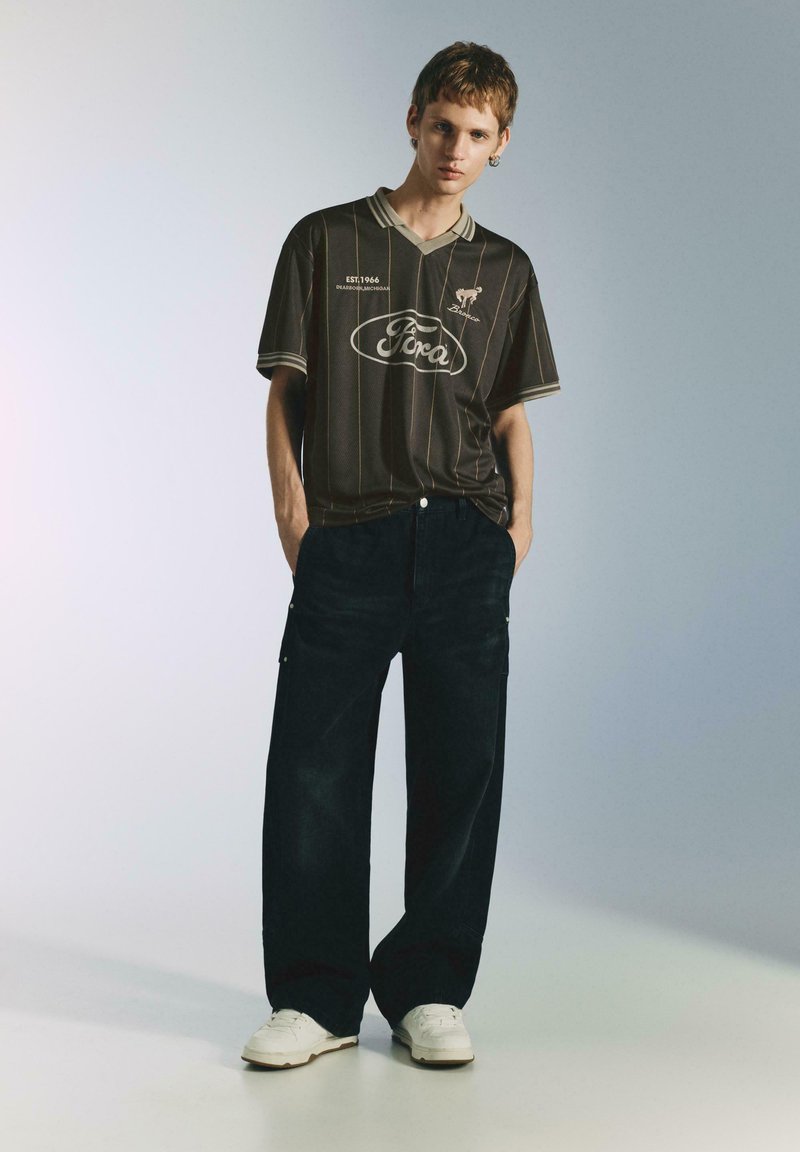 Polo a righe marroni con logo, indossata con jeans ampi scuri e sneakers bianche. Design semplice con dettaglio classico del colletto.