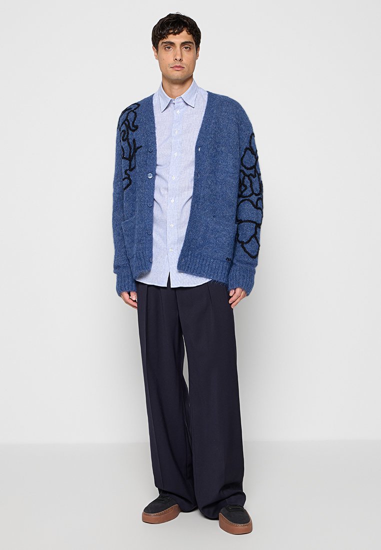 Hombre vestido con camisa azul abotonada, cárdigan azul oversize con patrón negro, pantalones anchos azul marino y zapatos negros con suelas marrones.