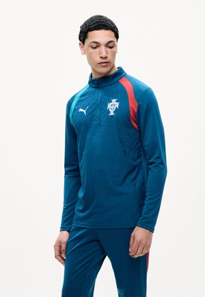 Muž oblečený v modrej športovej teplákovej súprave s červenými a tyrkysovými pruhmi a portugalským futbalovým erbom, pozerajúci sa dole do strany.