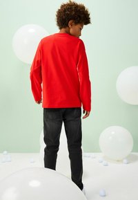 Red long-sleeve shirt, black jeans, standing on white floor with clear balloons. Background is light green. Focus on casual wear details.
