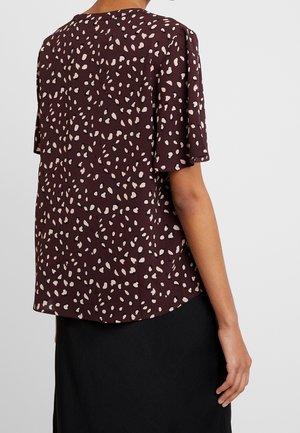 Kvinde iført en lilla kortærmet bluse med beige uregelmæssige pletter, kombineret med et sort skørt, vist bagfra.