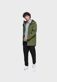 Grüne wasserdichte Jacke mit Kapuze, Frontreißverschluss und seitlichen Taschen, getragen über einem hellgrauen Hemd und kombiniert mit schwarzen Hosen und weißen Sneakern.