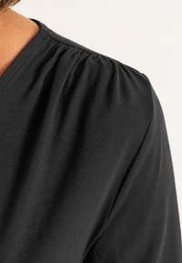 Schwarzes T-Shirt mit V-Ausschnitt und rüschendetail an der Schulter, gefertigt aus weichem Stoff, der eine glatte Textur und eine lockere Passform zeigt.