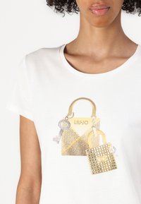 T-shirt bianco con una grafica di due borse metalliche dorate con dettagli in strass, una con una chiave e elementi texture.