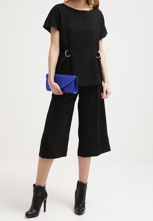 Zwarte korte mouw top met cirkelvormige accenten, gecombineerd met zwarte wijde culottes. Houdt een blauwe clutch vast en draagt zwarte enkelboots.