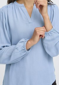 Femme ajustant le poignet élastique d'une blouse à manches longues bleu clair avec un col en V et des manches froncées.