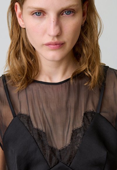 Mujer joven con piel clara, pecas y cabello castaño rojizo ondulado hasta los hombros, vestida con una blusa de encaje negro transparente sobre una camiseta negra.