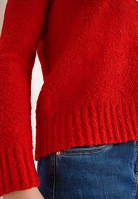 Červený, texturovaný svetr s dlouhými rukávy a ribbovaným lemem, spárovaný s modrými džínami. Svetr má načechraný, měkký materiál.