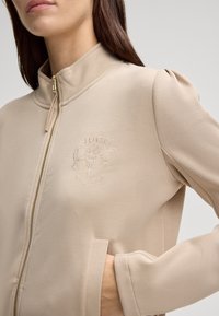 Giacca beige con zip e colletto alto, caratterizzata da un dettaglio del logo ricamato sul petto, una texture liscia e tasche laterali.