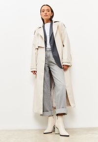 Kermainen trench coat, jossa on suuret napit, kerroksittain harmaan bleiserin ja valkoisen paidan päällä, yhdistettynä vaaleanharmaisiin farkkuihin ja nilkkureihin, joissa on tekstuuriset korkot.