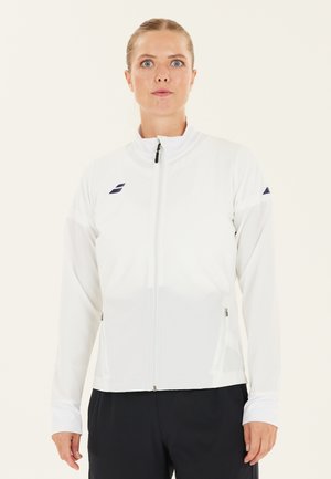 Kvinde med håret samlet i nakken iført en hvid jakke med lynlås og sorte logoer, stående foran en ensfarvet lys baggrund.