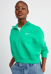 Sweatshirt verde com fecho éclair, feito de tecido suave, com punhos e barra canelados; apresenta o logótipo branco da Nike no lado esquerdo do peito. Usado com calças de ganga.