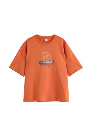 Smilende dreng iført en orange "Xtreme Communication" T-shirt, blå beanie, blå jeans og sorte sko, stående mod en ensfarvet lys baggrund.