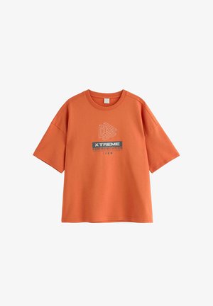 Smilende dreng iført en orange "Xtreme Communication" T-shirt, blå beanie, blå jeans og sorte sko, stående mod en ensfarvet lys baggrund.