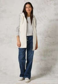 Lichtgrijze vest met capuchon en voorrits, geweven fleece voorkant, gebreide mouwen, gecombineerd met blauwe wijde jeans en witte sneakers.