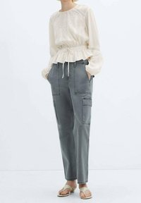 Blusa beige chiaro a maniche lunghe con vita elastica e cuciture decorative, abbinata a pantaloni cargo grigi con due tasche laterali e una vestibilità rilassata.