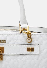 Weiße, strukturierte Handtasche mit goldenen Beschlägen, Reißverschluss und einem kleinen runden goldenen Anhänger mit einem Fragezeichen in der Nähe der Griffbefestigung.