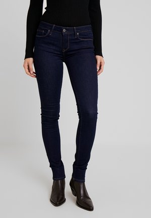 Donna con jeans skinny blu scuro, top nero a maniche lunghe a coste e stivaletti neri con punta a punta su uno sfondo grigio semplice.