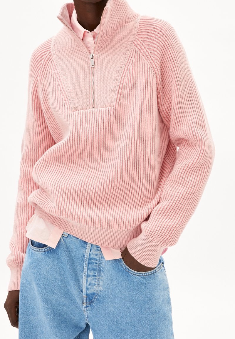 Personne portant un pull tricoté côtelé rose clair avec un col demi-fermeture éclair, par-dessus une chemise rose pâle et un jean bleu clair, les mains dans les poches.