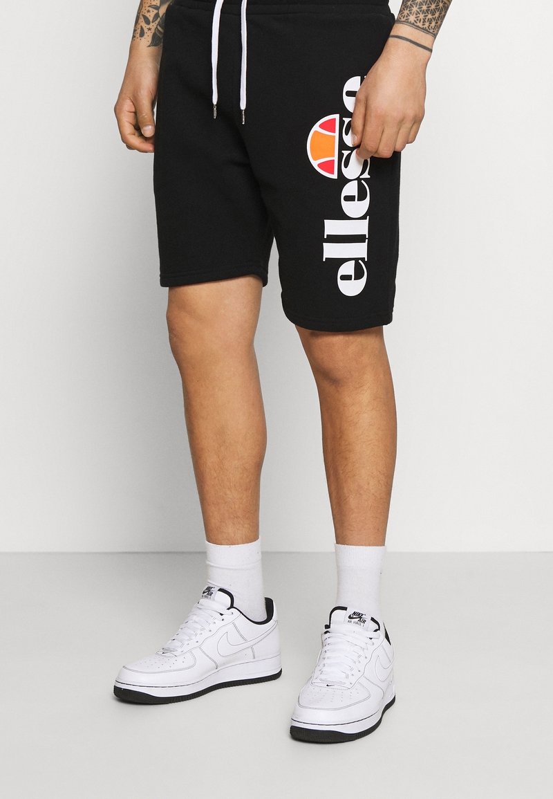 Personne portant un short noir Ellesse avec des cordons blancs, des baskets blanches Nike Air, des chaussettes blanches et des tatouages visibles sur l'avant-bras.