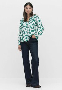 Vit långärmad skjorta med grönt abstrakt mönster av blad; avslappnad passform, krage och lätt tyg. Matchad med mörka utsvängda jeans.