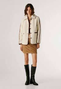 Krēmkrāsas šerpas jaka ar brūnām detaļām, pogām un apkakli. Kombinēta ar bēša krāsas svārkiem un melnām ceļgalu augstuma zābakiem.