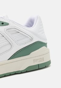 Baskets en cuir blanc avec des accents verts, présentant des panneaux texturés, une semelle en caoutchouc et un logo visible sur le côté.