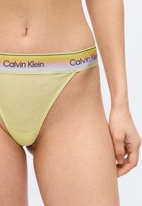 Κοντινό πλάνο ενός ατόμου που φοράει εσώρουχα Calvin Klein ανοιχτού πράσινου χρώματος με πολύχρωμη ζώνη μέσης σε αποχρώσεις του ουράνιου τόξου και μαύρο λογότυπο.